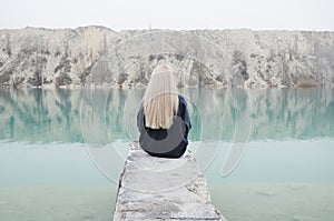 A girl with long blond hair sits at the end of the pier against the backdrop of a blue lake and hills
