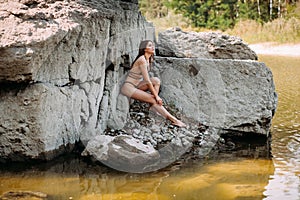 Girl with long blond hair, a slender figure, in a black swimsuit, sits on the rocks near the ocean