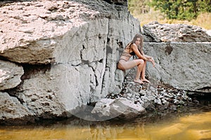 Girl with long blond hair, a slender figure, in a black swimsuit, sits on the rocks near the ocean