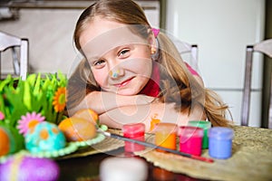 A girl with long blond ponytails and a painted nose sits at the kitchen table in front of decorated Easter eggs