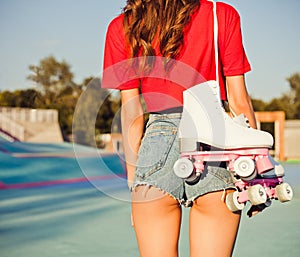 Girl with long dark hair is back with white roller skates on her shoulder. Warm summer evening in the skate park. Outdoor. Close