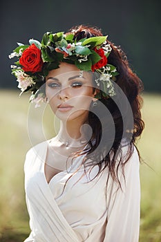 Girl in a long dress stands in a field with wreath on her head and bouquet of flowers in her hands, beautiful woman in the rays of