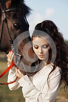 Girl in a long dress stands near a horse, a beautiful woman strokes a horse and holds the bridle in a field in autumn. Country