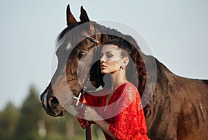 Girl in a long dress stands near a horse, a beautiful woman strokes a horse and holds the bridle in a field in autumn. Country