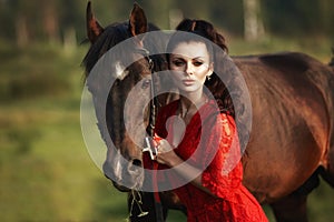 Girl in a long dress stands near a horse, a beautiful woman strokes a horse and holds the bridle in a field in autumn. Country