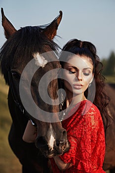 Girl in a long dress stands near a horse, a beautiful woman strokes a horse and holds the bridle in a field in autumn. Country