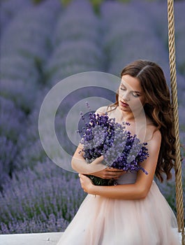 A girl with long flowing hair sits on a rope swing in a lavender field in a fluffy tulle dress at dawn with fog