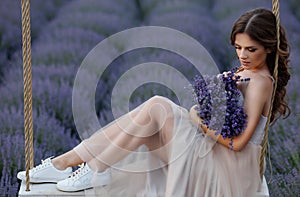 A girl with long flowing hair sits on a rope swing in a lavender field in a fluffy tulle dress at dawn with fog