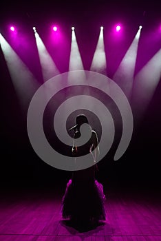 Girl in long gown performing on stage. Girl singing on the stage in front of the lights. Silhouette of singer standing