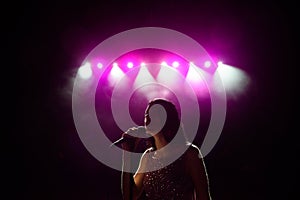 Girl in long gown performing on stage. Girl singing on the stage in front of the lights. Silhouette of singer standing