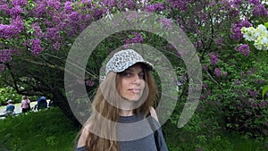 A Girl with long hair in a baseball cap stands in front of a blooming lilac.