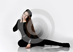 Girl with long hair, in black blouse, beret, pants, high heel boots. She smiling, sitting on floor, isolated on white. Close up