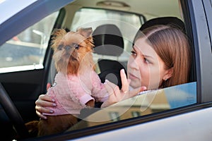 Girl with long hair in a black jacket and a small dog in pink clothes near window of a car