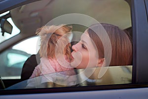 Girl with long hair in a black jacket and a small dog in pink clothes near window of a car