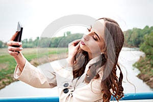 Girl with long hair and curls in a dress and coat standing on suspension bridge makes selfie