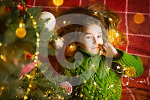 A girl with long hair and garlands lies on a red plaid under a Christmas tree with toys in a warm knitted sweater. Christmas, New