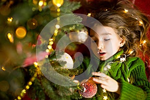 A girl with long hair and garlands lies on a red plaid under a Christmas tree with toys in a warm knitted sweater. Christmas, New