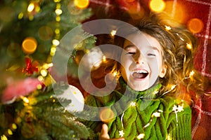 A girl with long hair and garlands lies on a red plaid under a Christmas tree with toys in a warm knitted sweater. Christmas, New