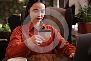 Girl with long hair holding credit card and using laptop making payment online sitting in cafe. Online shopping, e