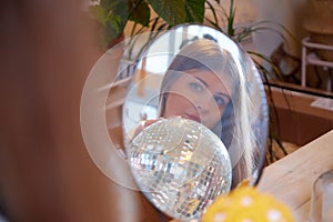 Girl with long hair looking on reflection in mirrow in a room with a lot of things. Portrait of a young woman with