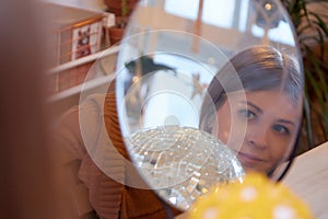 Girl with long hair looking on reflection in mirrow in a room with a lot of things. Portrait of a young woman with mirror in cozy