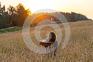 A girl with long hair with an old acoustic guitar stands in a wheat field lit by the rays of the setting sun and admires the