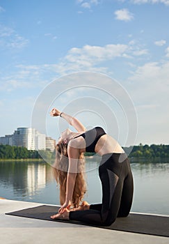 Girl with long hair practicing yoga outdoors near lake, sitting in Camel pose or Ustrasana.