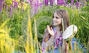 Girl with long hair sits in a clearing with lupine flowers and plays with a tambourine