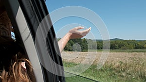 Girl with long hair is sitting in front seat of car, stretching her arm out window .