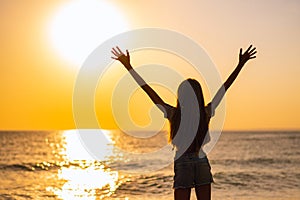 A girl with long hair stands on the seashore with her hands up.