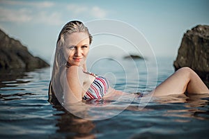 Girl with long hair, in a striped swimsuit, by the ocean, sits by the rocks, in the water and smiles