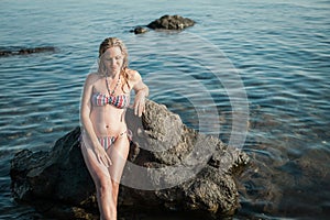 Girl with long hair, in a striped swimsuit, by the ocean, sits by the rocks, in the water and smiles
