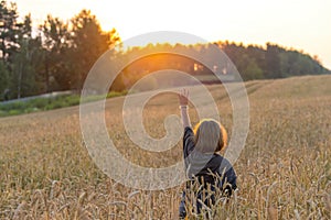 A girl with long hair in a wheat field lit by the rays of the sunset and greeting the sun by raising her hand