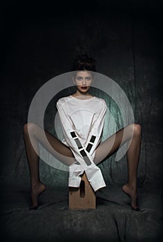 A girl with long legs in black tights and a white straitjacket sits on a wooden pedestal with her legs spread to the side photo