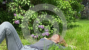 A girl with long red hair lies on the grass near a purple lilac bush.