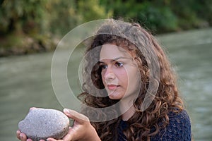 Girl with long wavy brown hair. With a stone in his hands along the bank of a river.