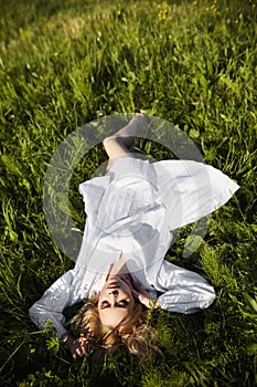Girl in a long white dress lies on the grass in a field. Blonde woman in the sun in a light dress. Girl resting and dreaming,