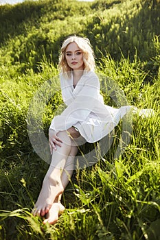 Girl in a long white dress sits on the grass in a field. Blonde woman in the sun in a light dress. Girl resting and dreaming,