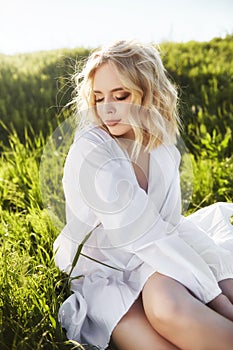 Girl in a long white dress sits on the grass in a field. Blonde woman in the sun in a light dress. Girl resting and dreaming,