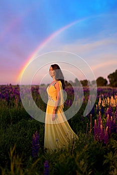 A girl in a long yellow dress against the background of a blooming purple lupine field and a sunset sky.