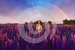 A girl in a long yellow dress against the background of a blooming purple lupine field and a sunset sky.