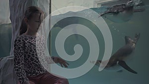 Girl look through glass in oceanarium at swimming penguins