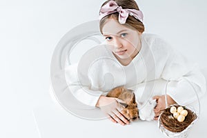 Girl looking at camera while sitting with furry rabbits and basket with easter eggs in nest on white