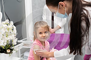 The girl is looking at the camera. A young lady doctor is preparing a child for an injection by disinfecting a piece of