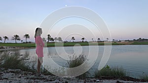 The girl is looking at game on golf course at dusk. Woman stand close to pond under the moon, dreams and thinks. Buggy