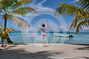 A girl looking horizon standing next to a palm trees on a beach in front of a Caribbean sea