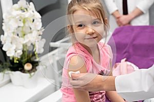 The girl is looking outside the frame. A young lady doctor is preparing a child for an injection by disinfecting a piece