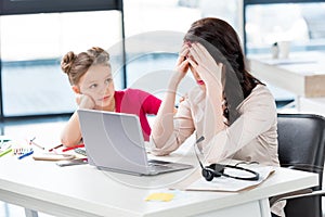 Girl looking at stressed mother working with laptop in office