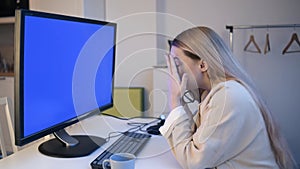 The girl looks into the green screen of the monitor. A beautiful blonde with glasses works at home in front of a large computer.