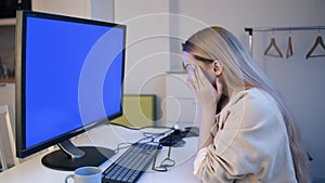 The girl looks into the green screen of the monitor. A beautiful blonde with glasses works at home in front of a large computer.